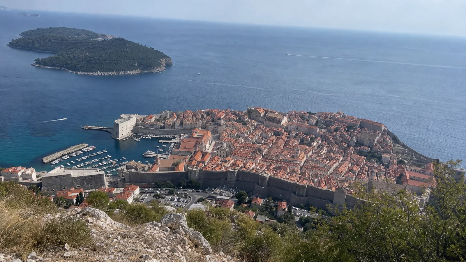 Elevated view of a walled coastal town with red roofs and offshore islands.
