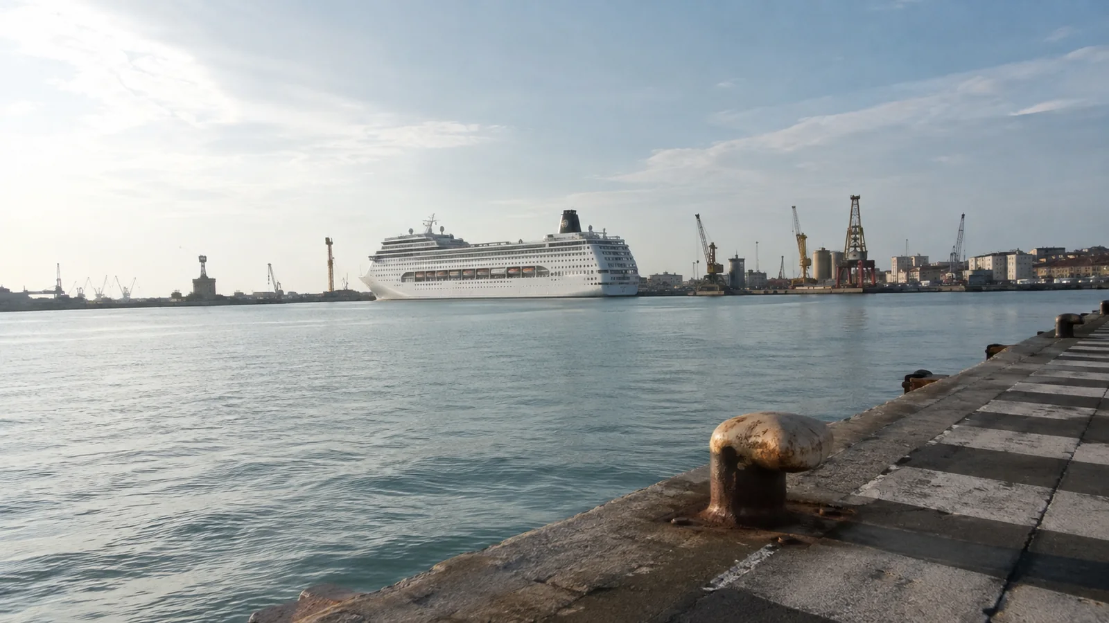 Cruise ship across an industrial harbor viewed from a stone quay.