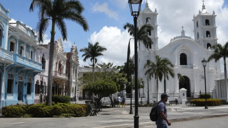 Palm-lined square with a white twin-towered church and colorful buildings.