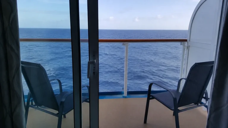 Two balcony chairs face the ocean beyond glass stateroom doors.