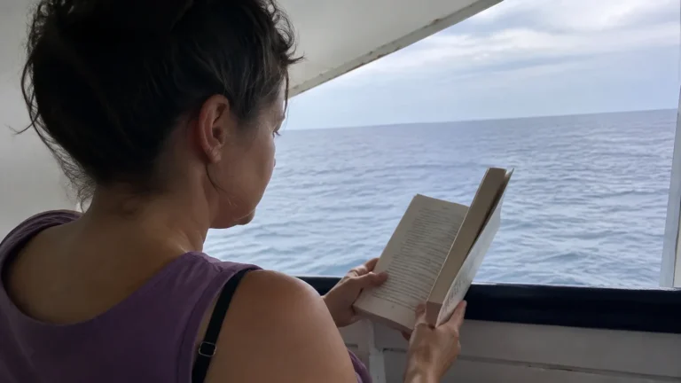 Passenger reading a paperback on a sheltered deck overlooking the ocean.