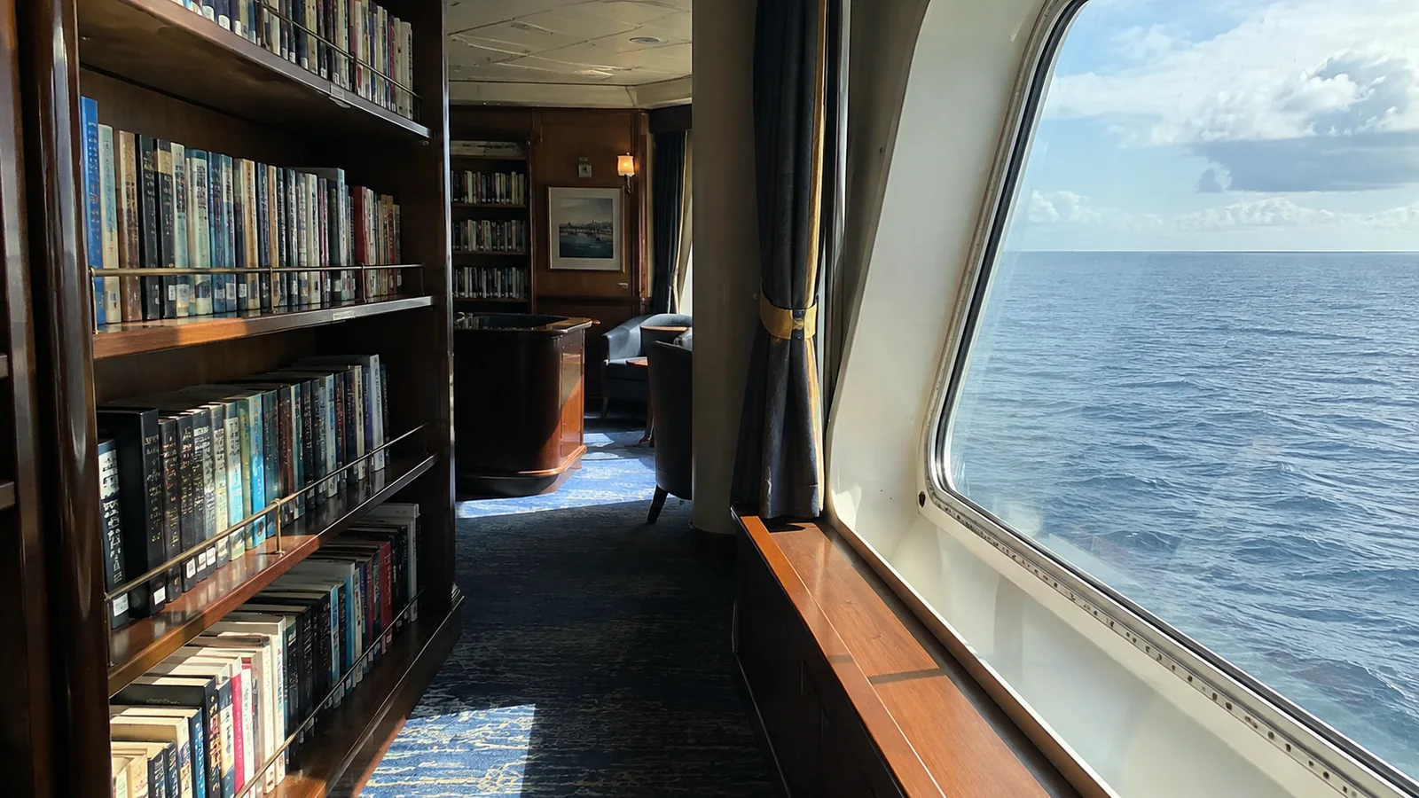 Bookshelves lining a quiet ship library beside large ocean-view windows.