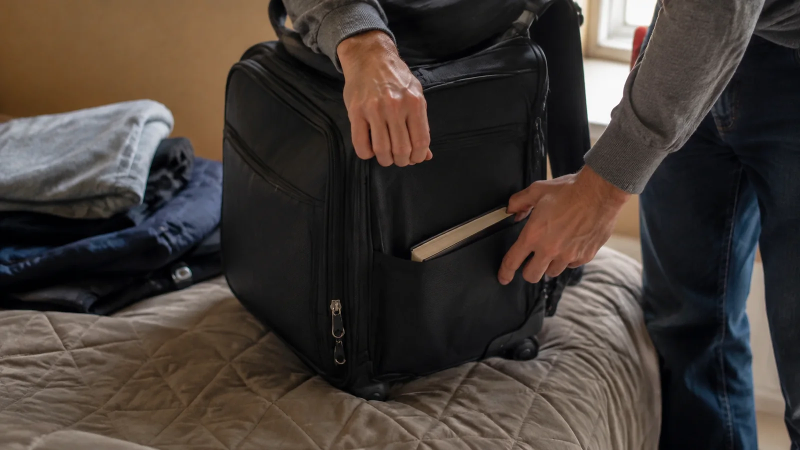 Traveler putting a book into the front pocket of a carry-on suitcase.