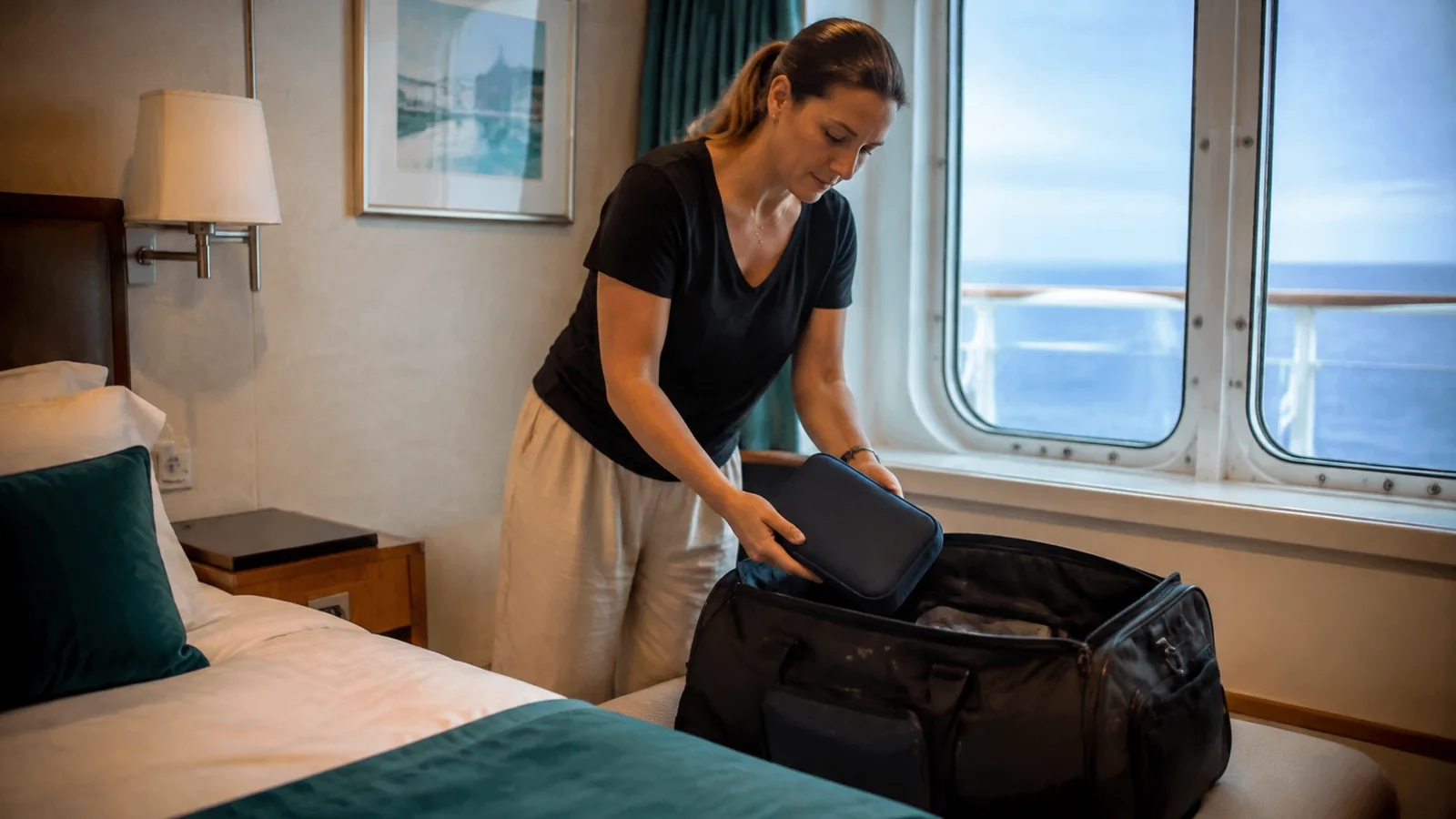 Woman packs a dark case into a bag beside ocean windows.