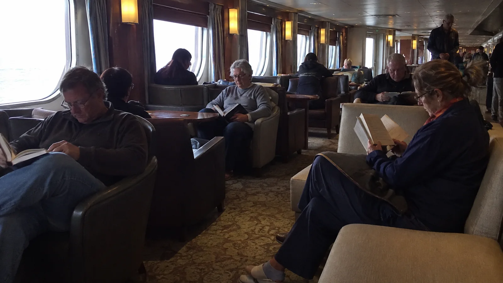 Adults reading quietly in an ocean-view ship lounge.