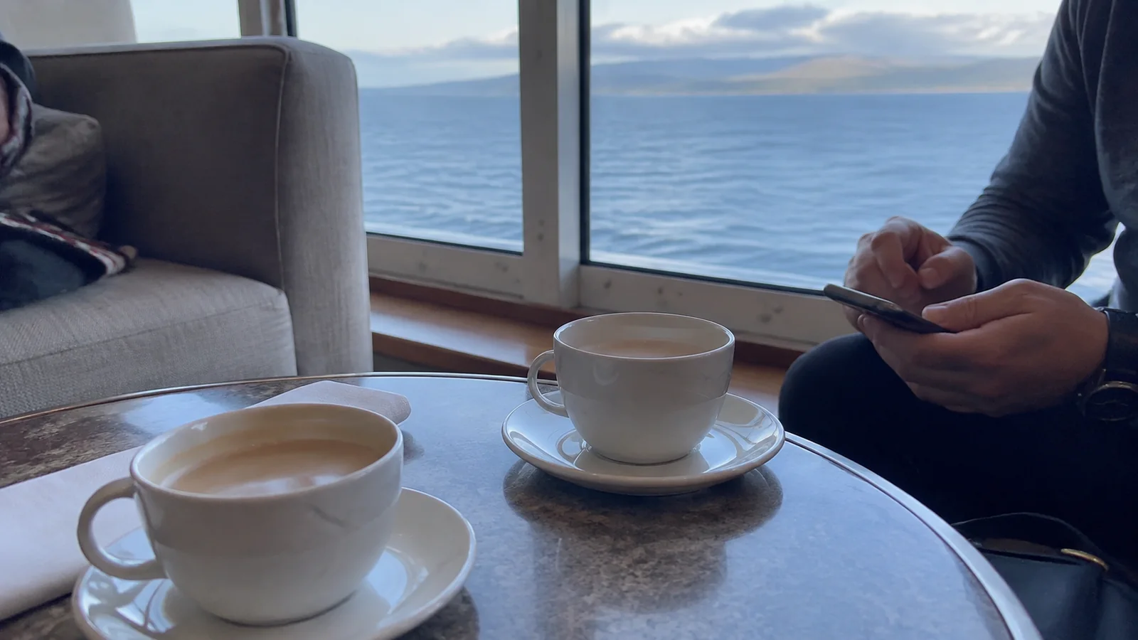 Two cups of coffee on a lounge table beside a passenger using a phone.