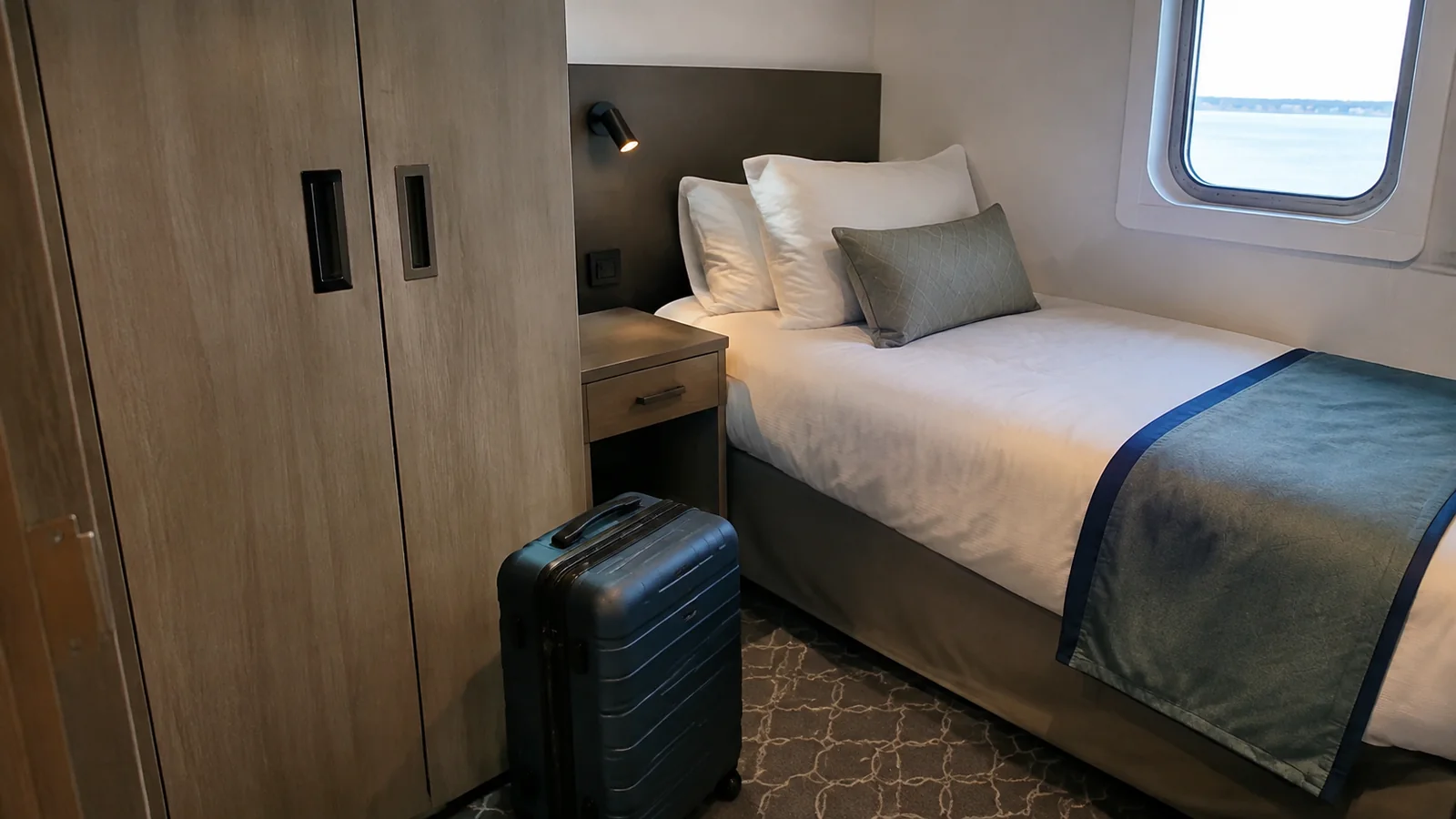 Single-bed cruise cabin with a window and small suitcase.