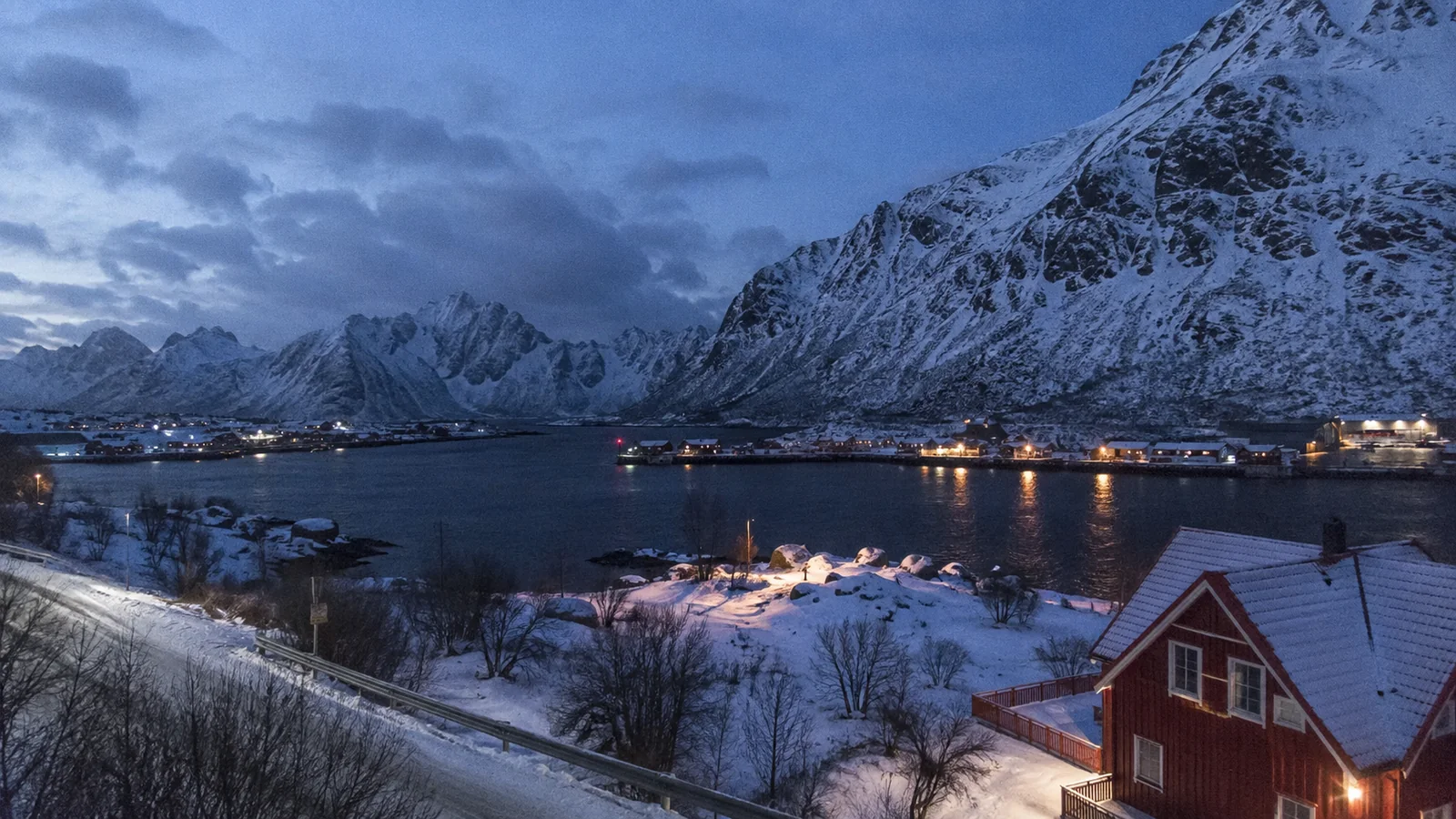 Snow-covered mountains and a waterfront village at blue hour.