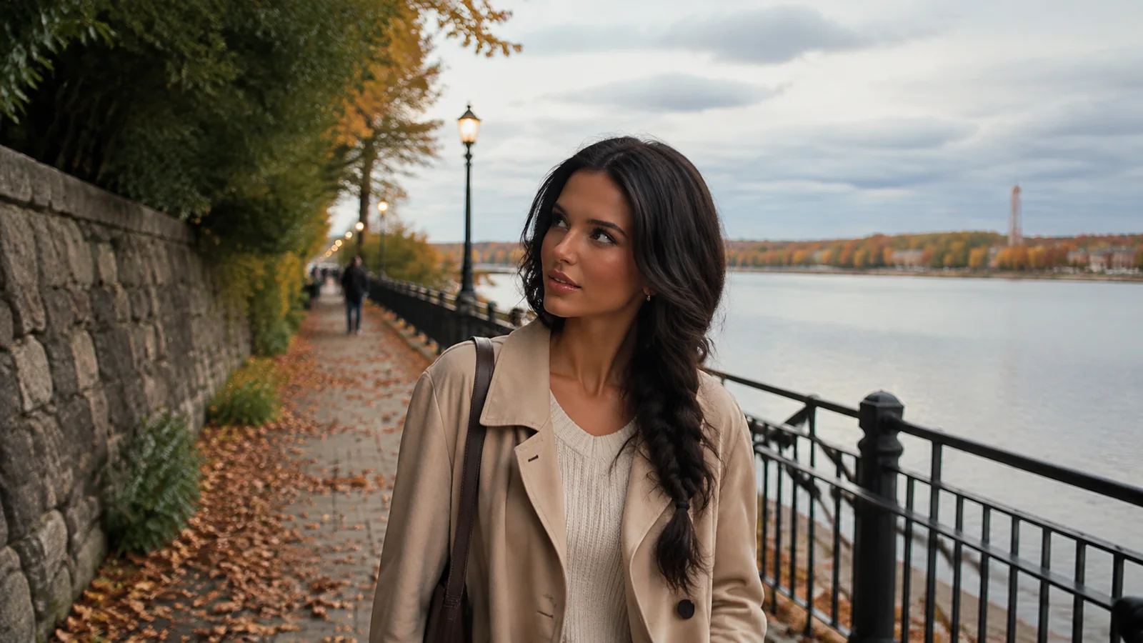 Woman in a trench coat walking along a leaf-strewn waterfront path.