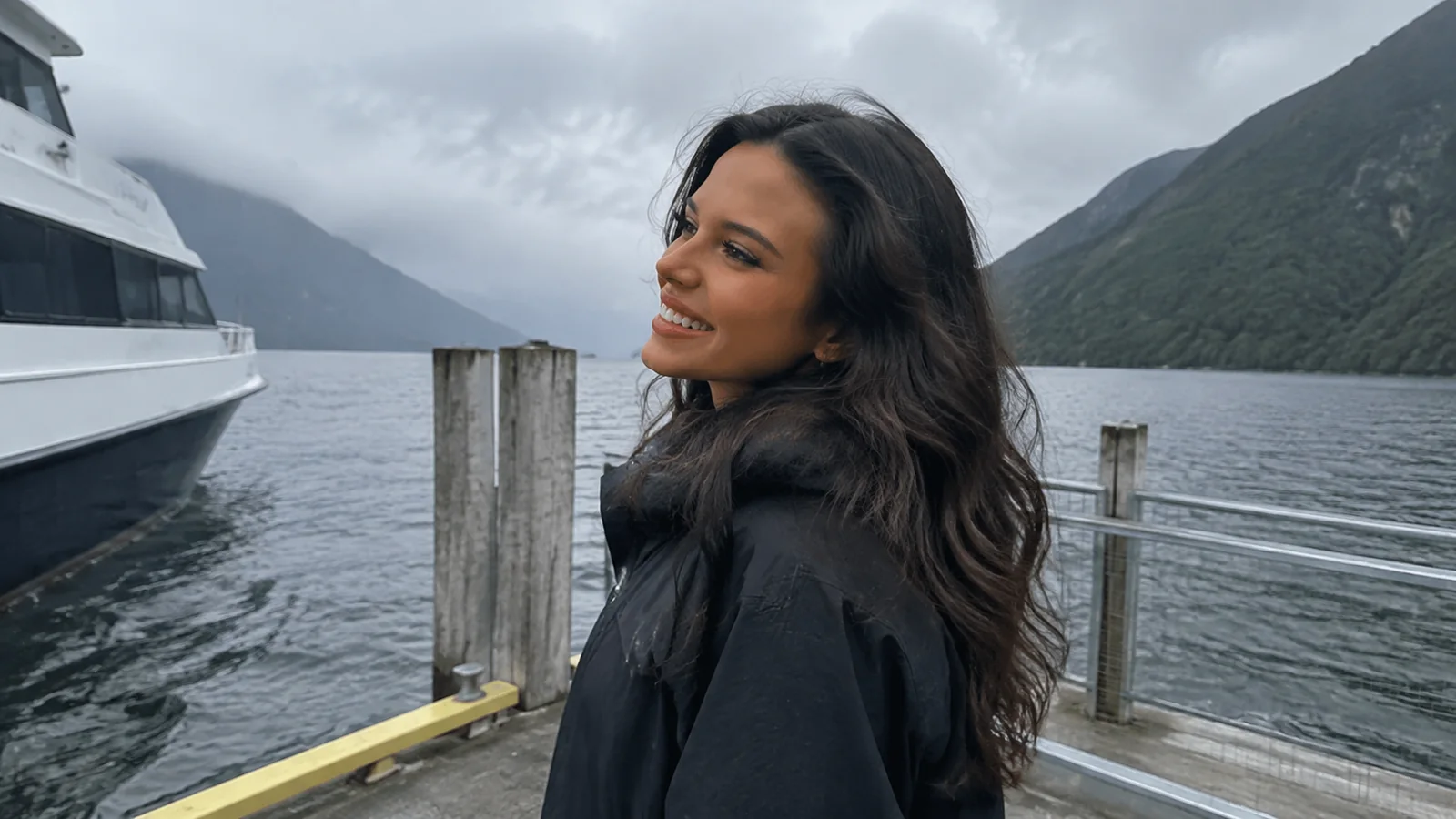 Woman standing on a jetty beside an excursion vessel and forested mountains.