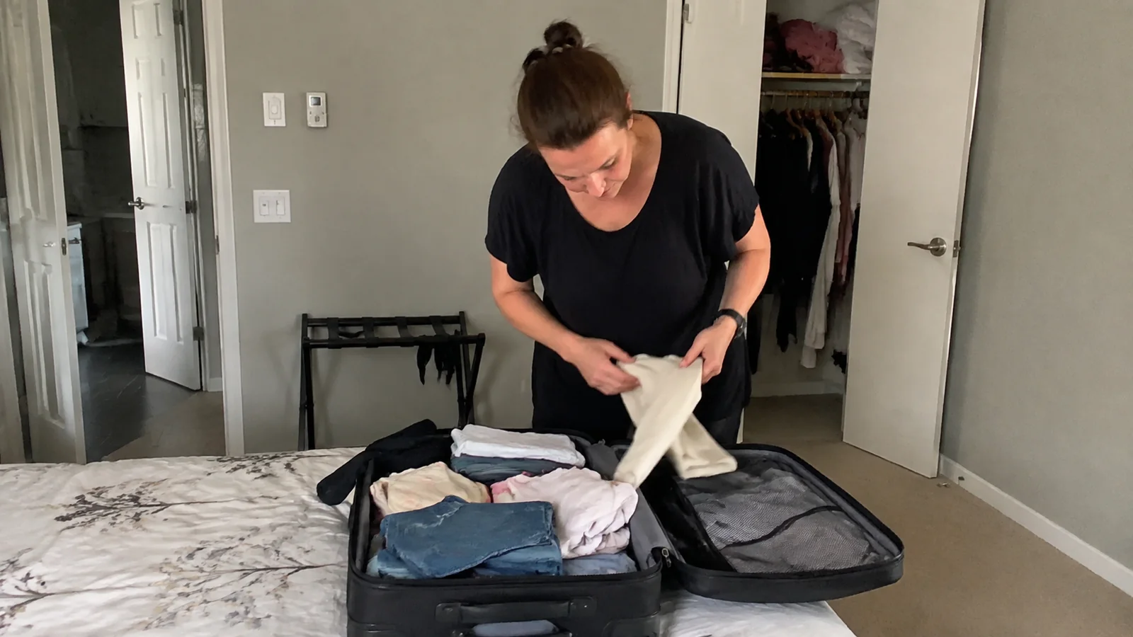 Woman folding white clothing into an open suitcase on a bed.
