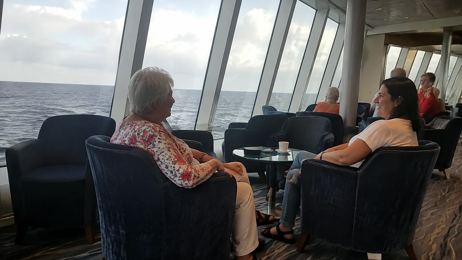 Two women chatting in lounge chairs beside large ocean-facing windows.
