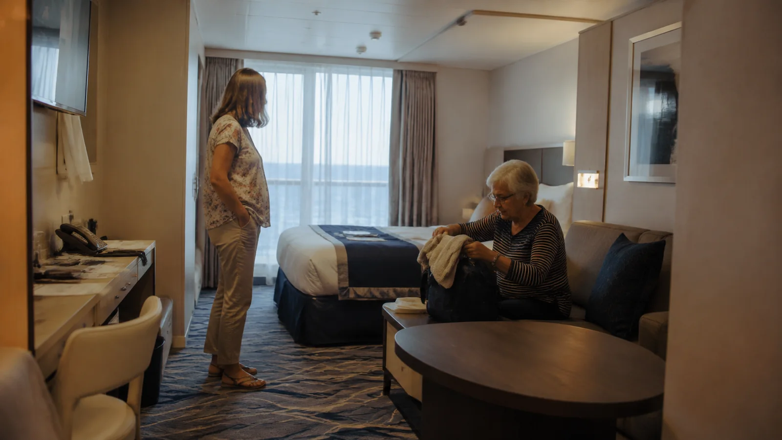 Two women talking inside a ship cabin with a bed and sofa.