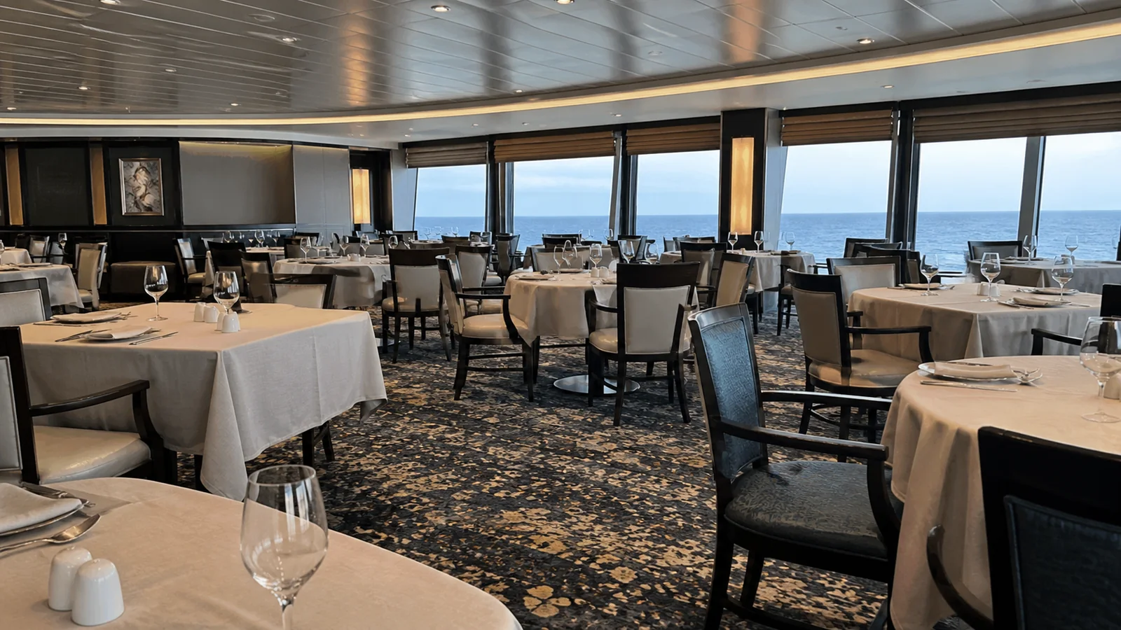 Empty ship restaurant with neatly set tables and ocean-facing windows.