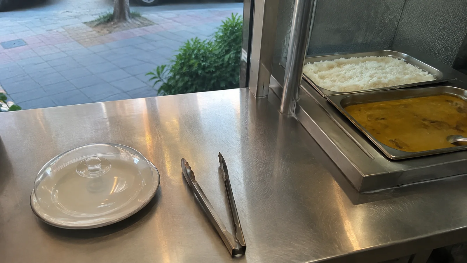 Covered empty plate and serving tongs beside open buffet trays of rice and curry.