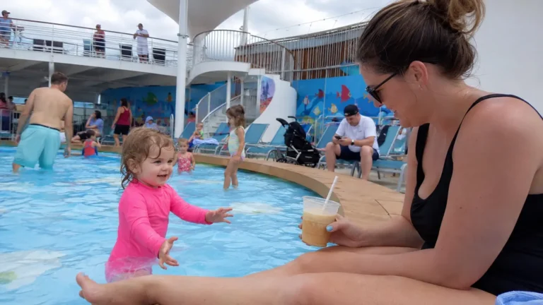 Woman sitting poolside beside a toddler in a pink swimsuit in a shallow pool