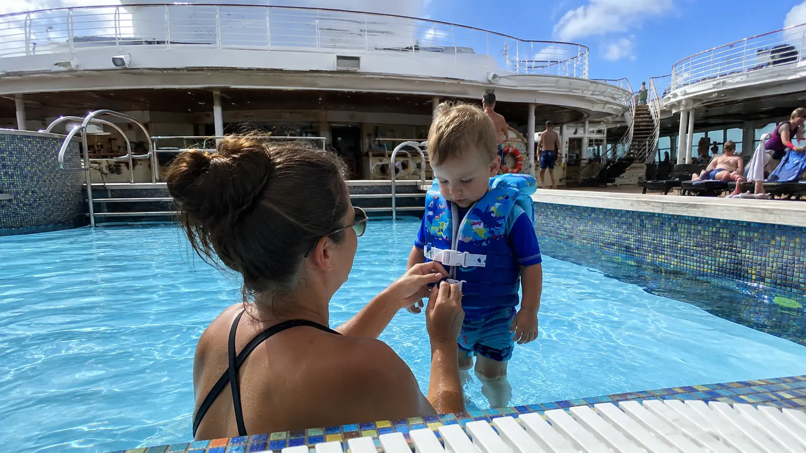 Woman helping a toddler wearing a flotation vest at the edge of a cruise ship pool