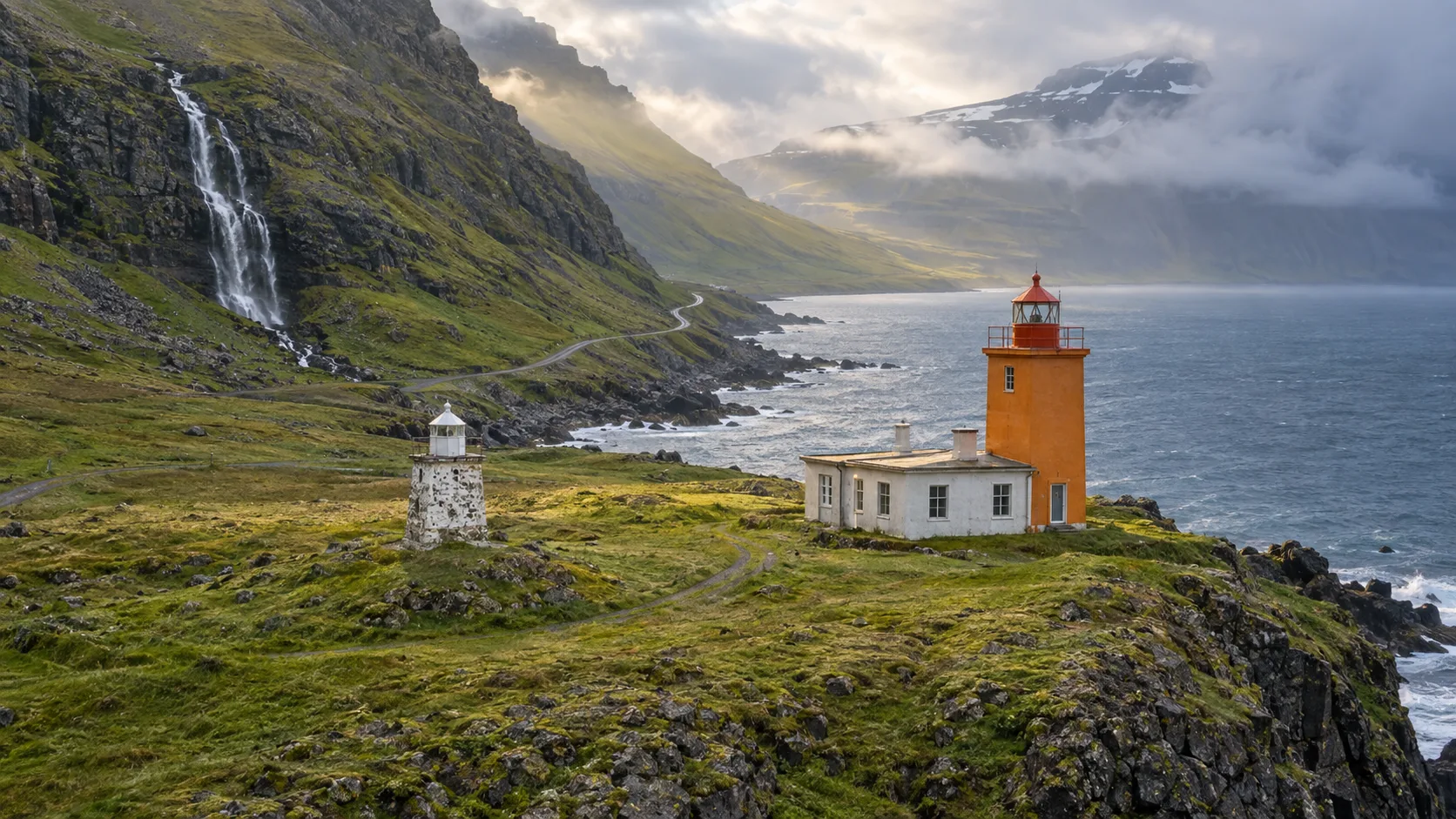 The old stone and active orange Dalatangi lighthouses beside a waterfall-lined East Iceland fjord.