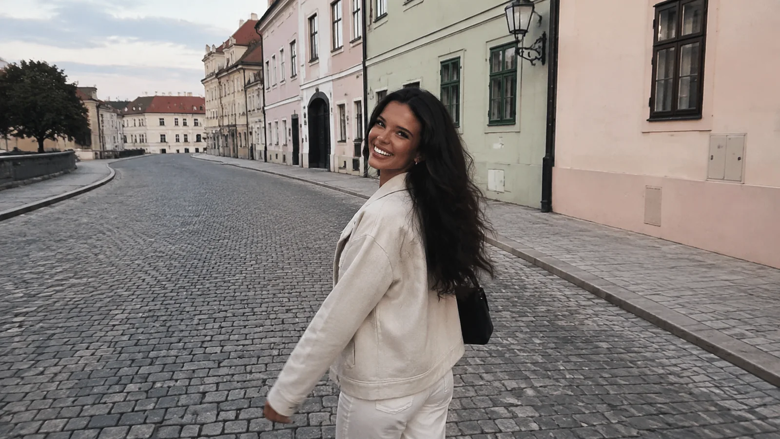 Woman walking along a cobblestone street lined with pale historic buildings.
