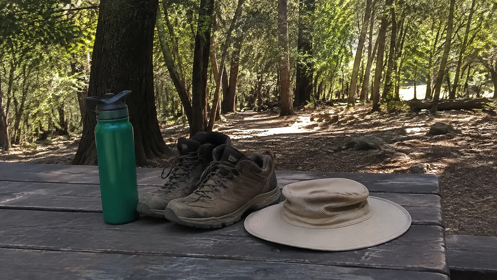 Hiking boots, water bottle, and sun hat rest on a woodland picnic table.