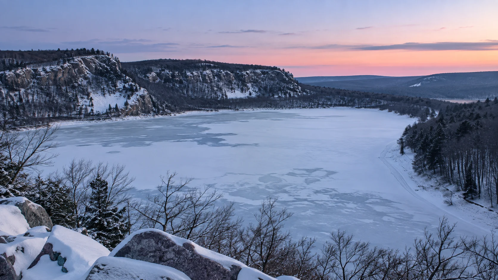 Snow-covered bluffs surround frozen Devil's Lake beneath a pale pink winter sky.
