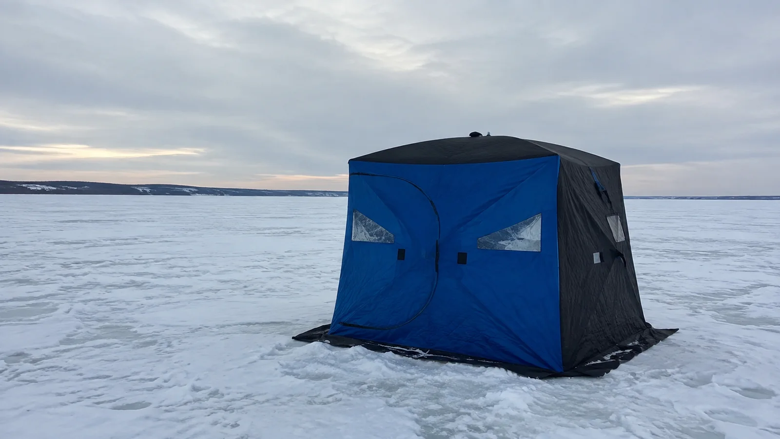 Blue ice fishing shelter stands alone on a snow-covered lake.