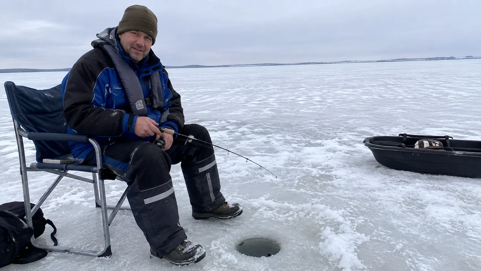 Bundled angler seated on a chair beside fishing gear on ice.