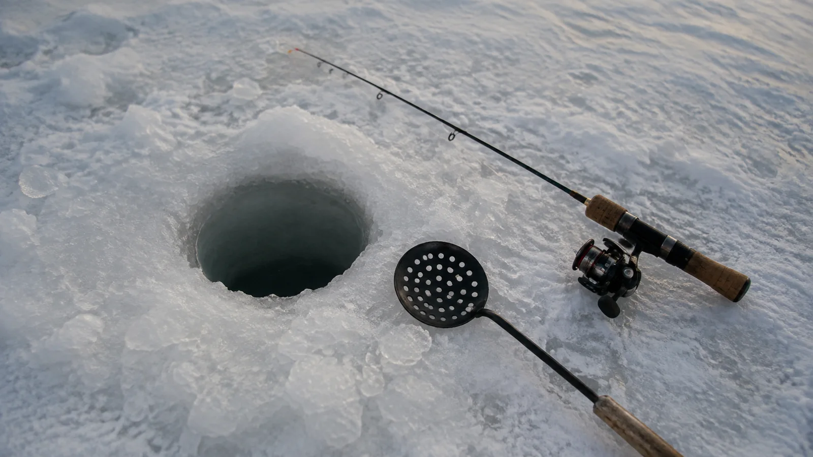 Fishing rod and ice scoop beside a round hole in the ice.