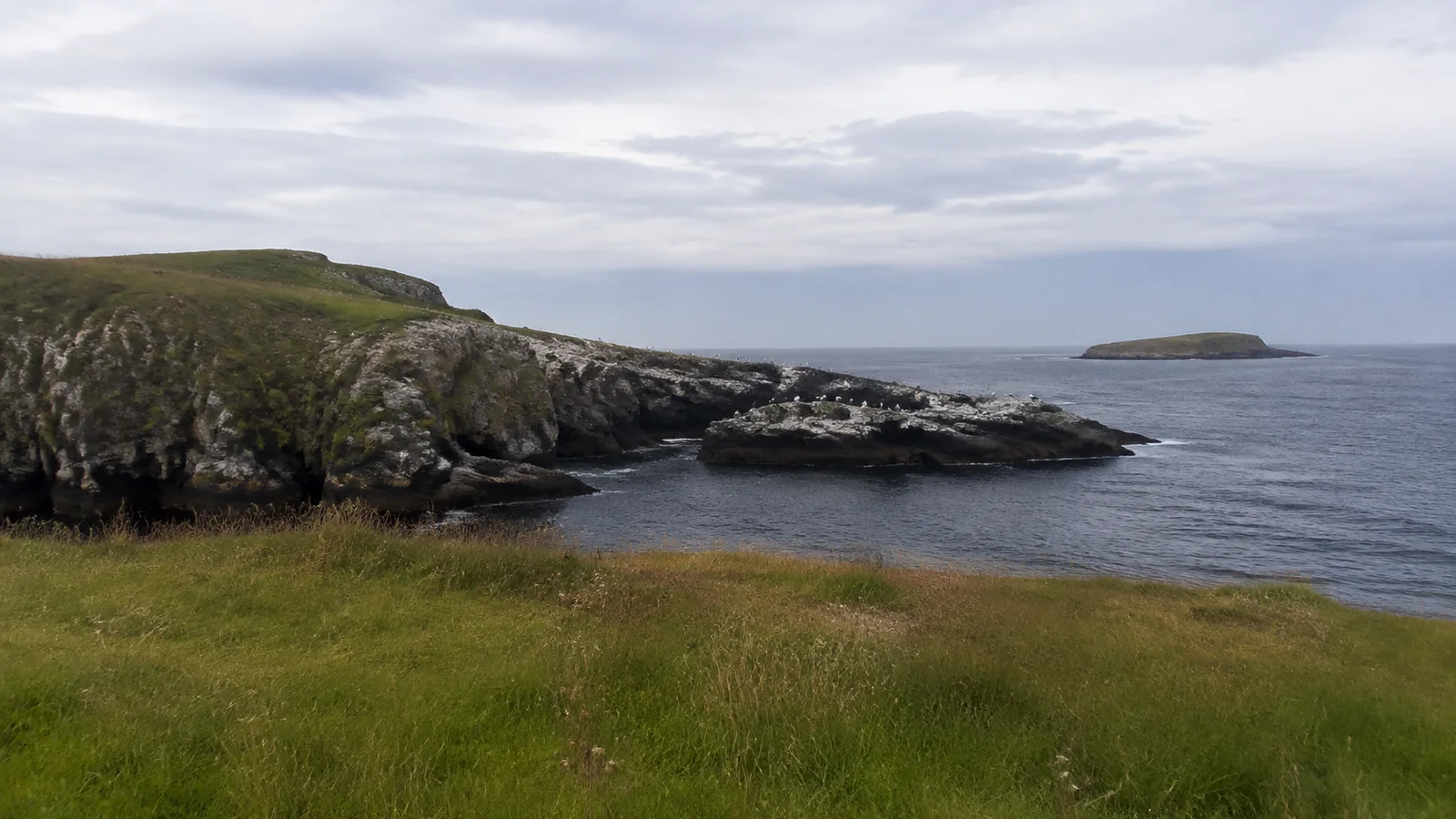 Rocky coastal inlet beneath grassy cliffs and cloudy skies.