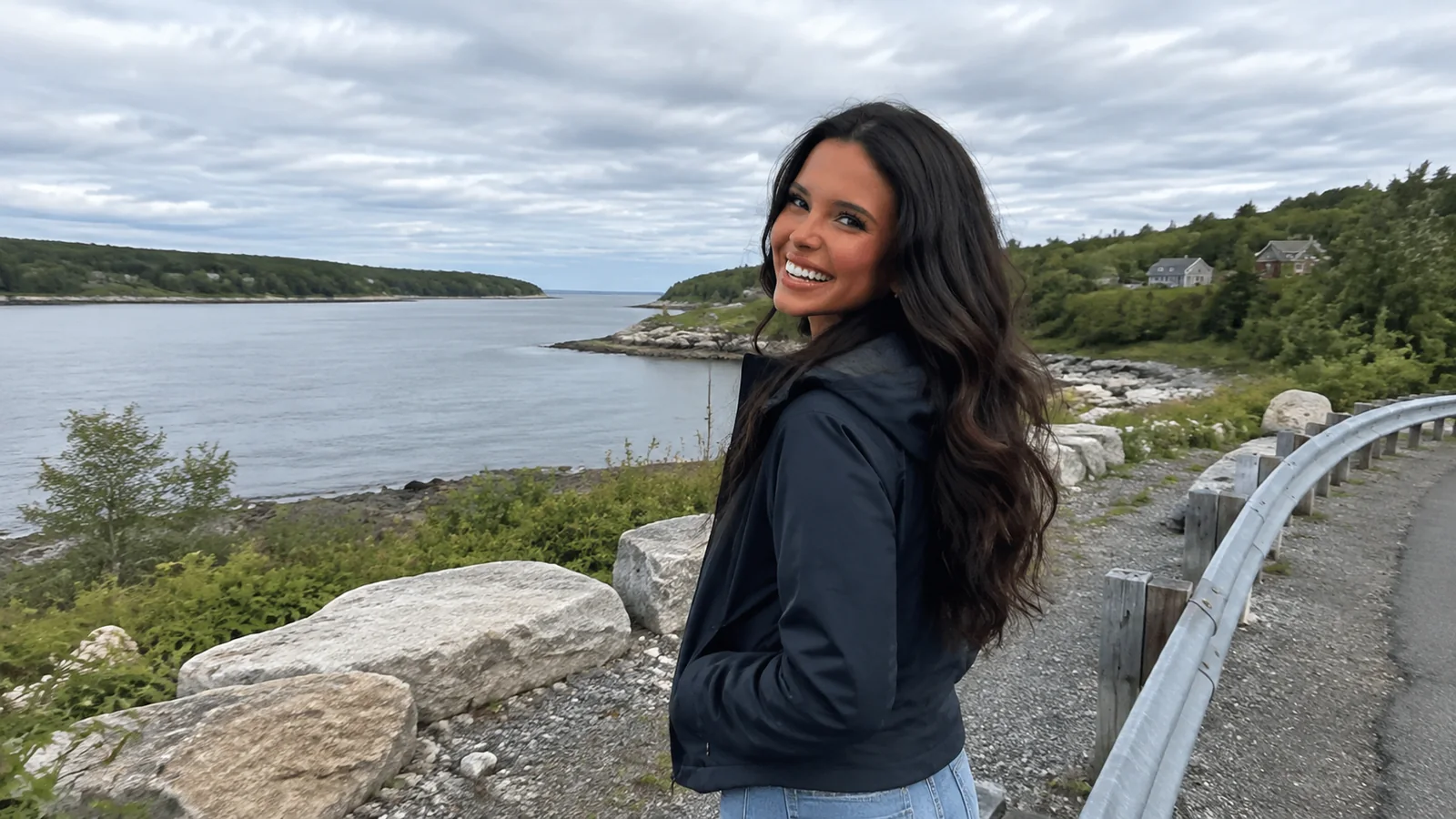 Woman standing beside a roadside railing overlooking a rocky inlet.