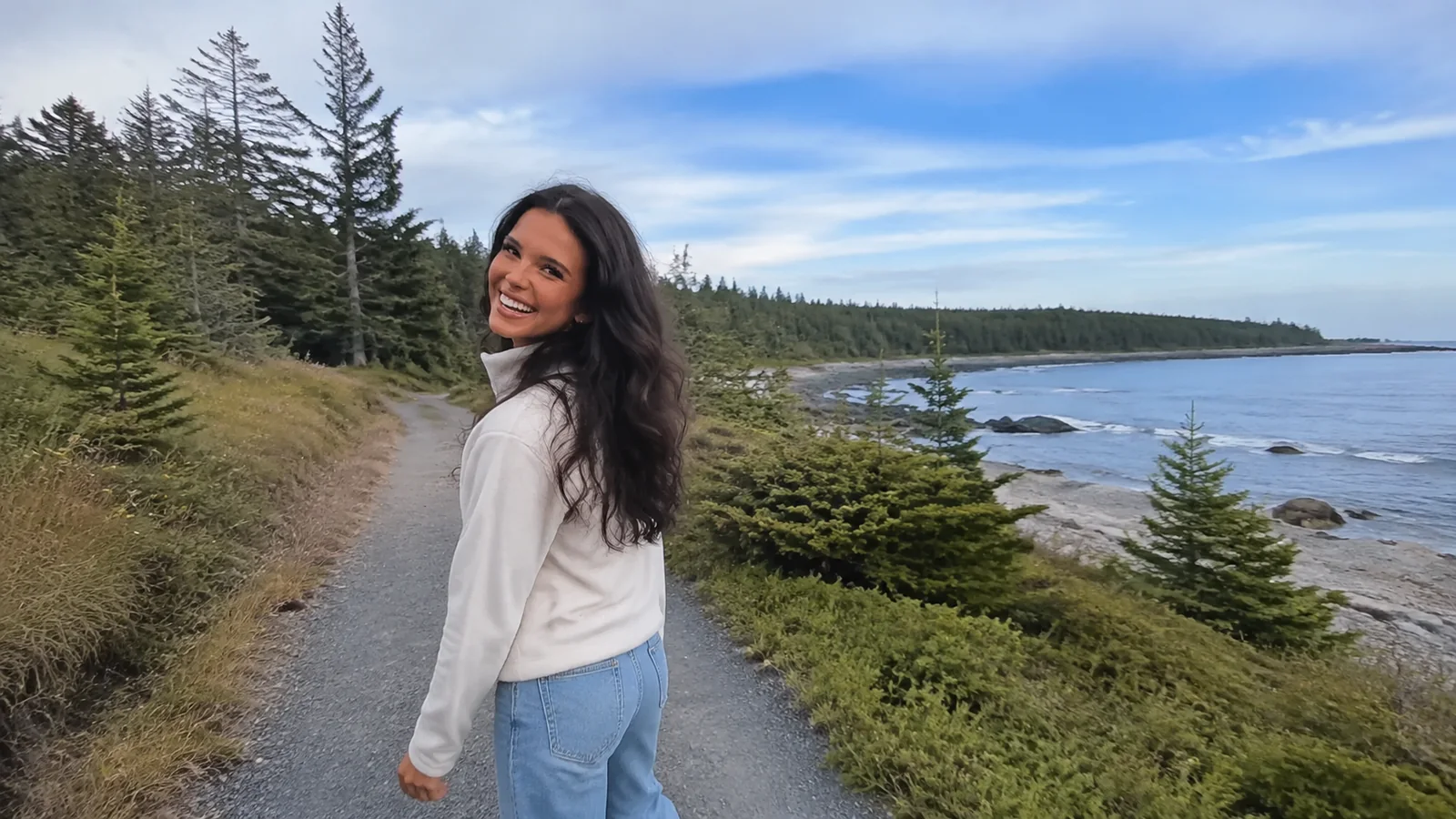 Woman walking a gravel coastal path beside low evergreens.