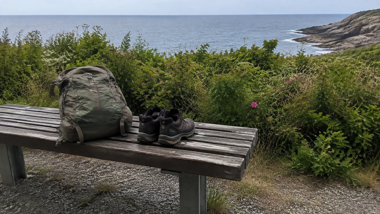 Backpack and hiking shoes resting on a bench above a rocky shoreline.