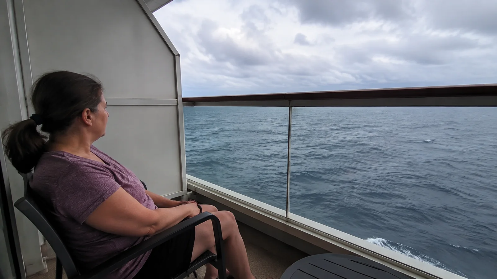 Passenger relaxing in a chair on a narrow ocean-facing balcony.