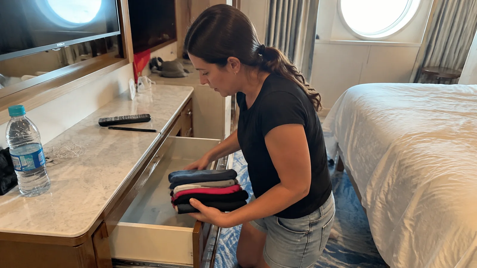 Woman places folded clothing into a drawer beside a stateroom bed.