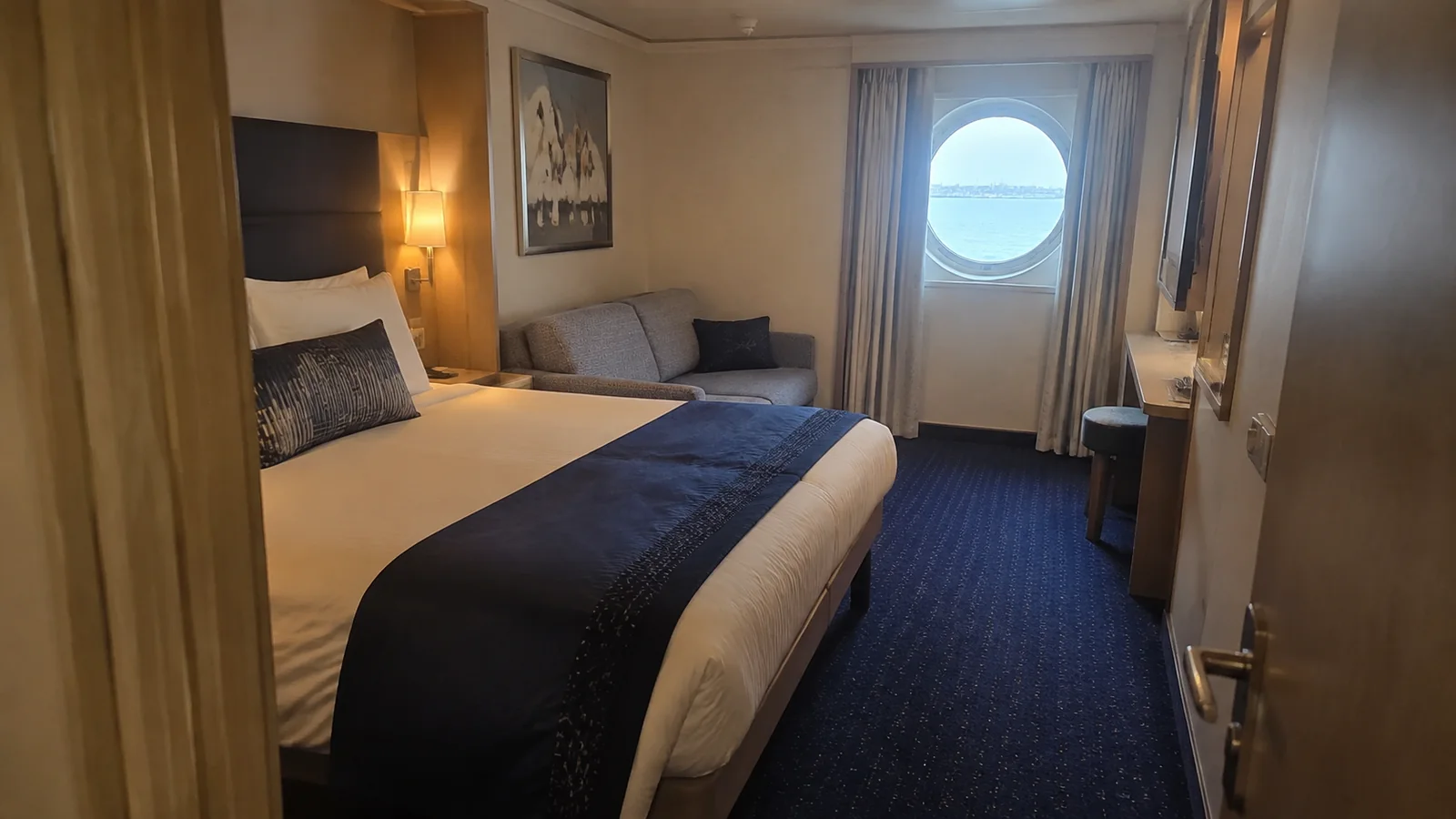 Compact stateroom with a double bed, sofa, blue carpet, and round ocean window.