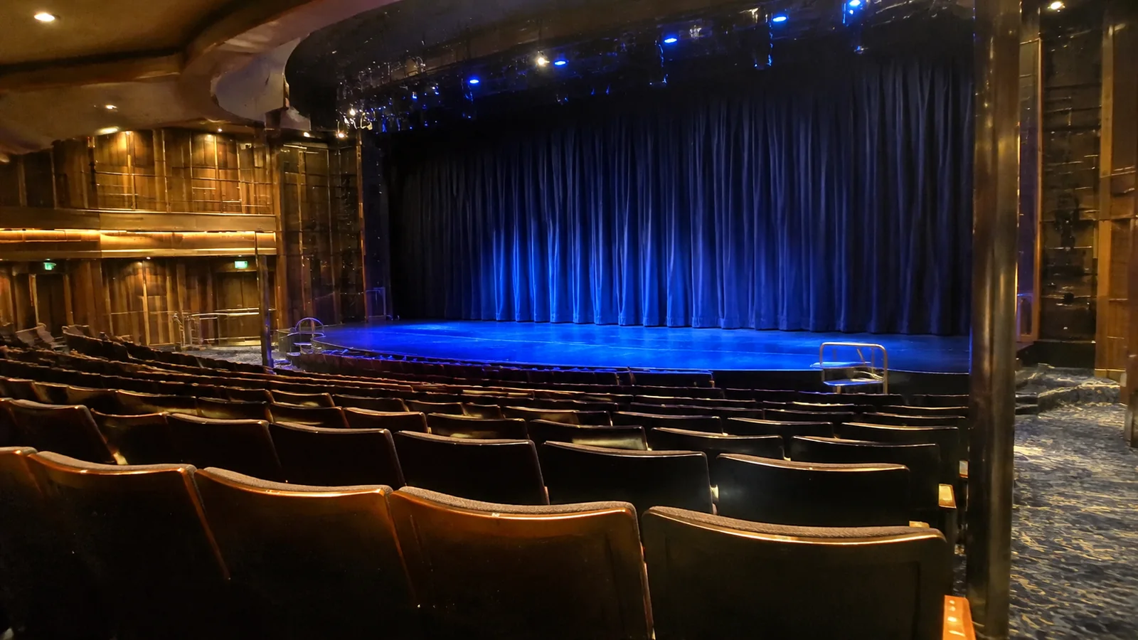 Empty theater with rows of seats facing a blue curtain and illuminated stage.