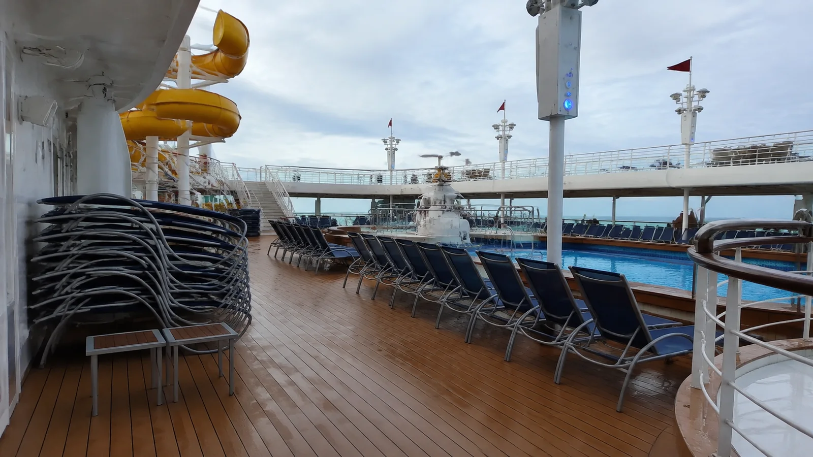 Outdoor ship deck with stacked loungers, a pool, and yellow waterslide.