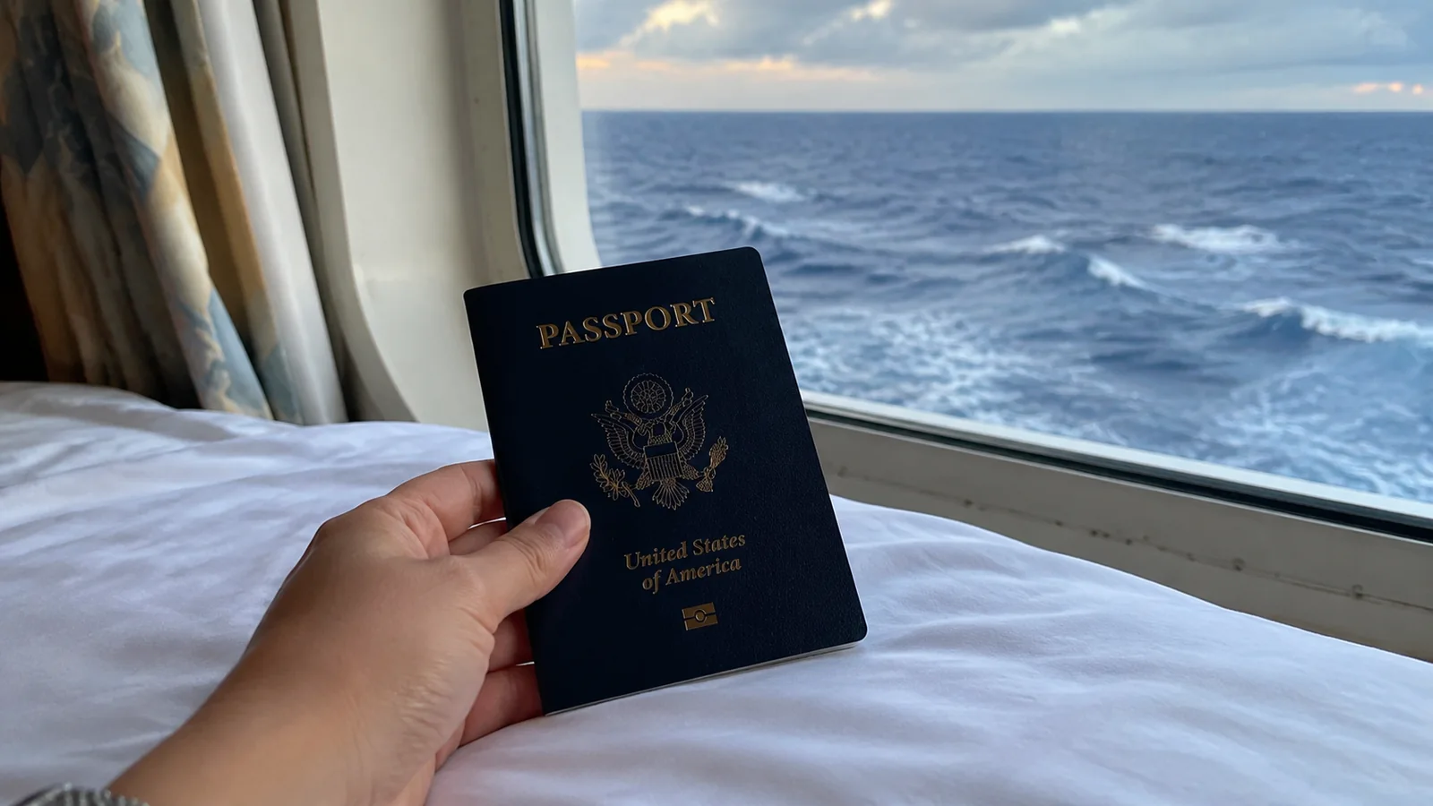 Hand holding a passport over a cruise cabin bed beside an ocean-view window