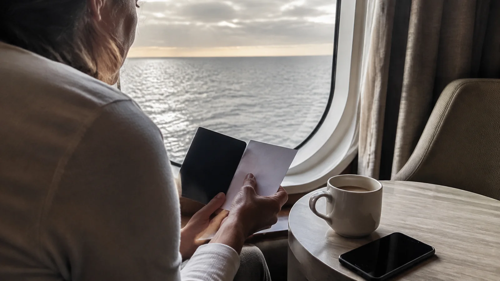 Person holding travel documents beside a cabin window and coffee cup