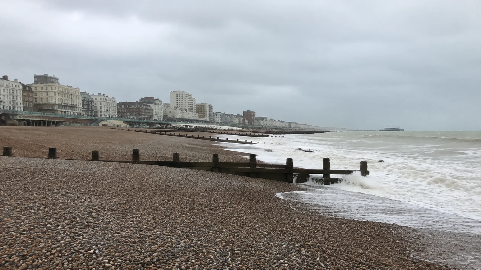 Wooden groynes stretch across a pebble beach toward gray water.