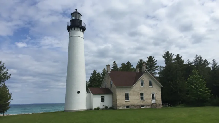 Tall white lighthouse and keeper's house stand beside a wooded shoreline.