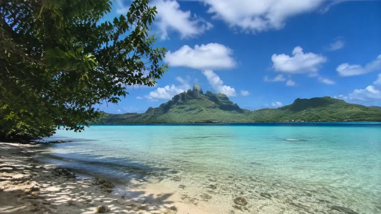 Clear turquoise lagoon beside a sandy shore with green volcanic peaks beyond.