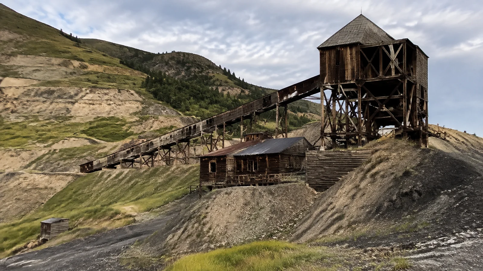 Weathered wooden mining structure and conveyor stand below barren hills.
