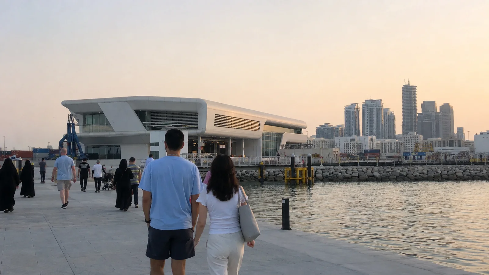 People walking along a waterfront toward a modern passenger terminal at dusk.