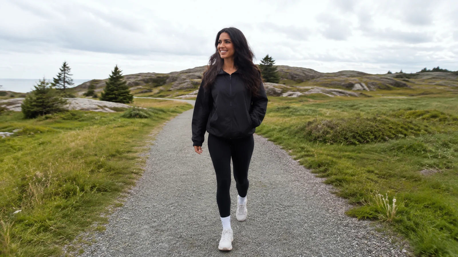 Woman walking a gravel path through open grassy coastal terrain.