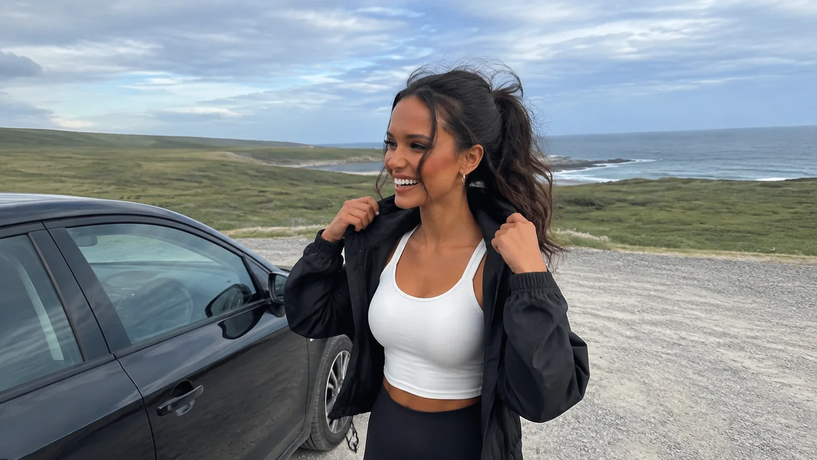 Woman adjusting her jacket beside a car on an open coast.