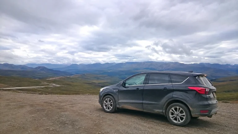 SUV parked at a gravel overlook above a broad mountain landscape.