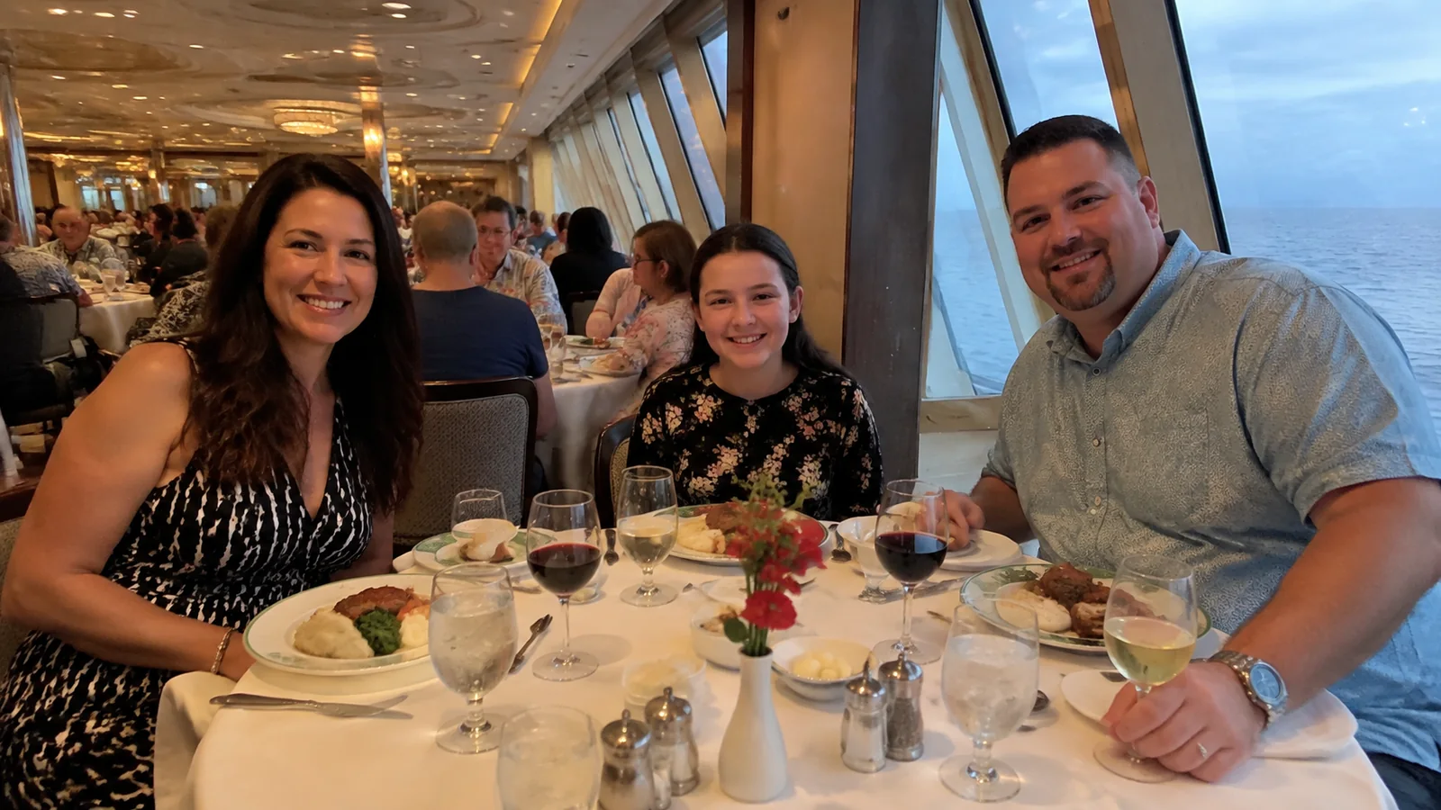 Three travelers smile around a dining table beside ocean windows.