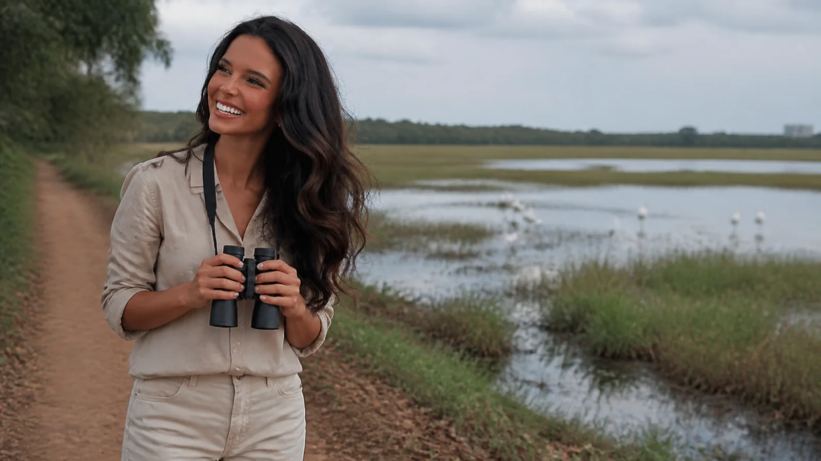 Woman holds binoculars on a dirt path beside wetland birds.