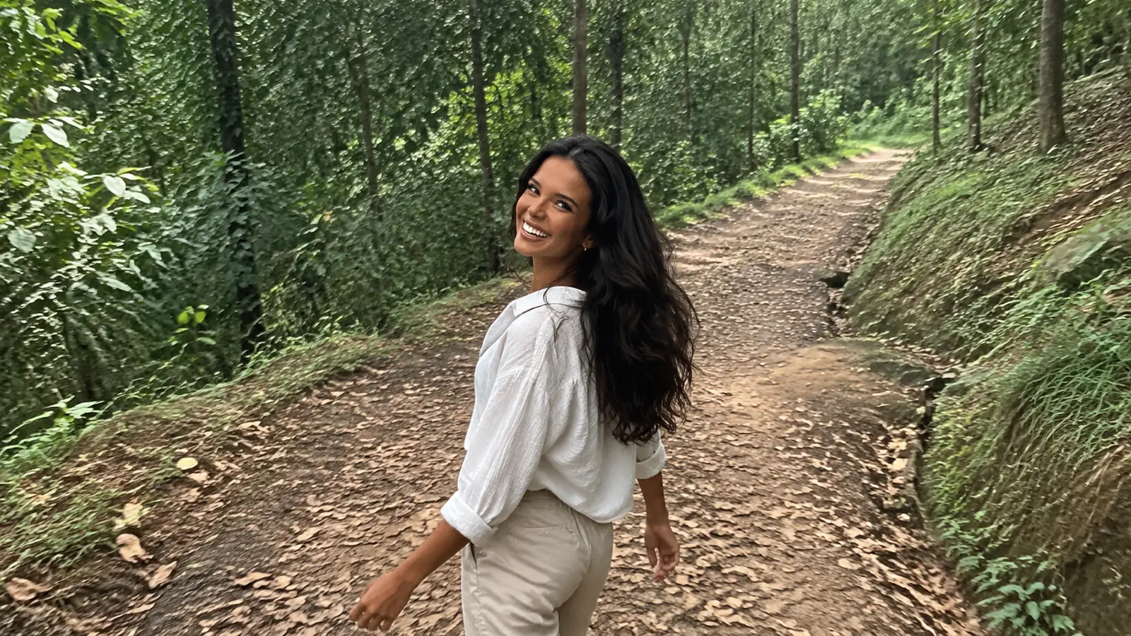 Woman walking a narrow green forest trail beneath leafy trees.